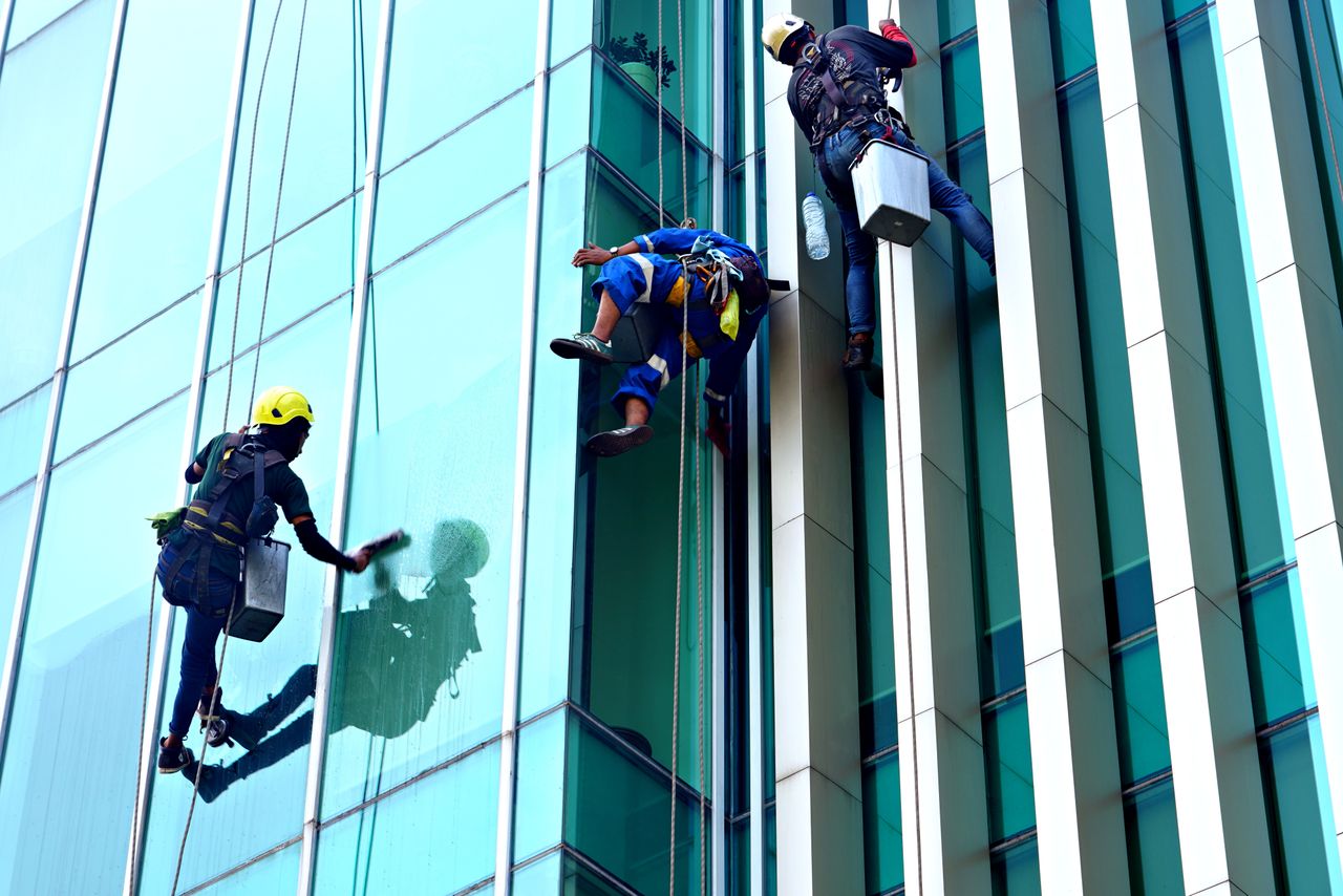 services-02 Three window cleaners hanging from ropes are cleaning a contemporary glass building facade in daylight.